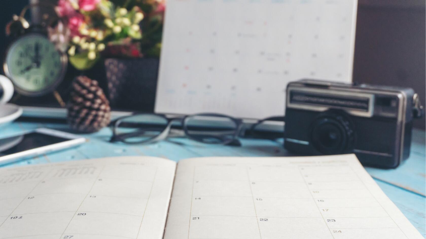 A calendar on a desk with a camera, glasses, and a cup of coffee