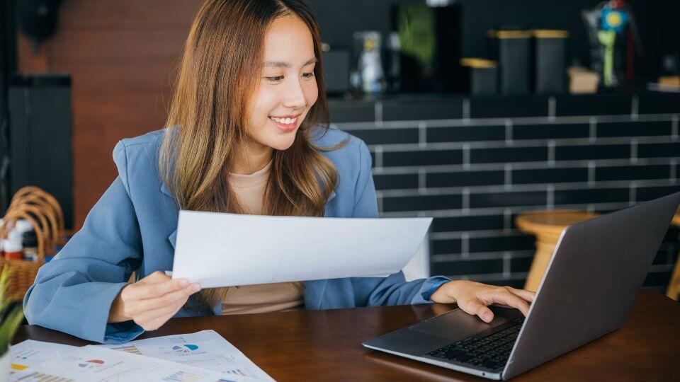 A woman holding a piece of paper with data on it and looking at her laptop