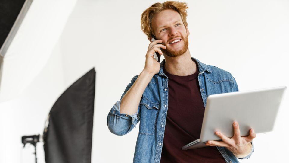 smiling man holding laptop and talking on cell phone in studio